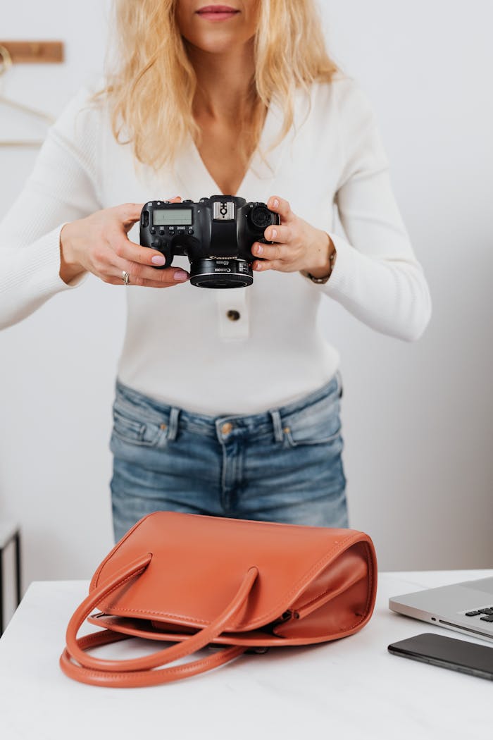 Blonde woman in white top taking photos of a handbag placed on a table.